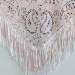 May include: A triangular bandana featuring a light pink background with a dark gray paisley and floral pattern. The edges of the fabric are finished with a long, white fringe trim that hangs down from the bottom point of the triangular shape.