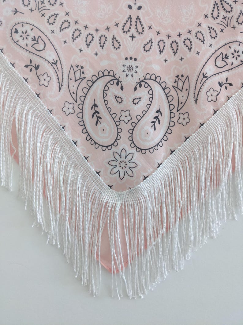 May include: A triangular bandana featuring a light pink background with a dark gray paisley and floral pattern. The edges of the fabric are finished with a long, white fringe trim that hangs down from the bottom point of the triangular shape.