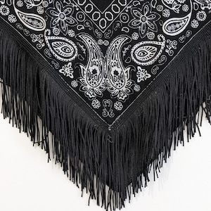 May include: A black triangular bandana featuring a white paisley and floral pattern printed across the fabric. The edges of the bandana are finished with long, thin black fringe that hangs down from the bottom point and sides of the garment.