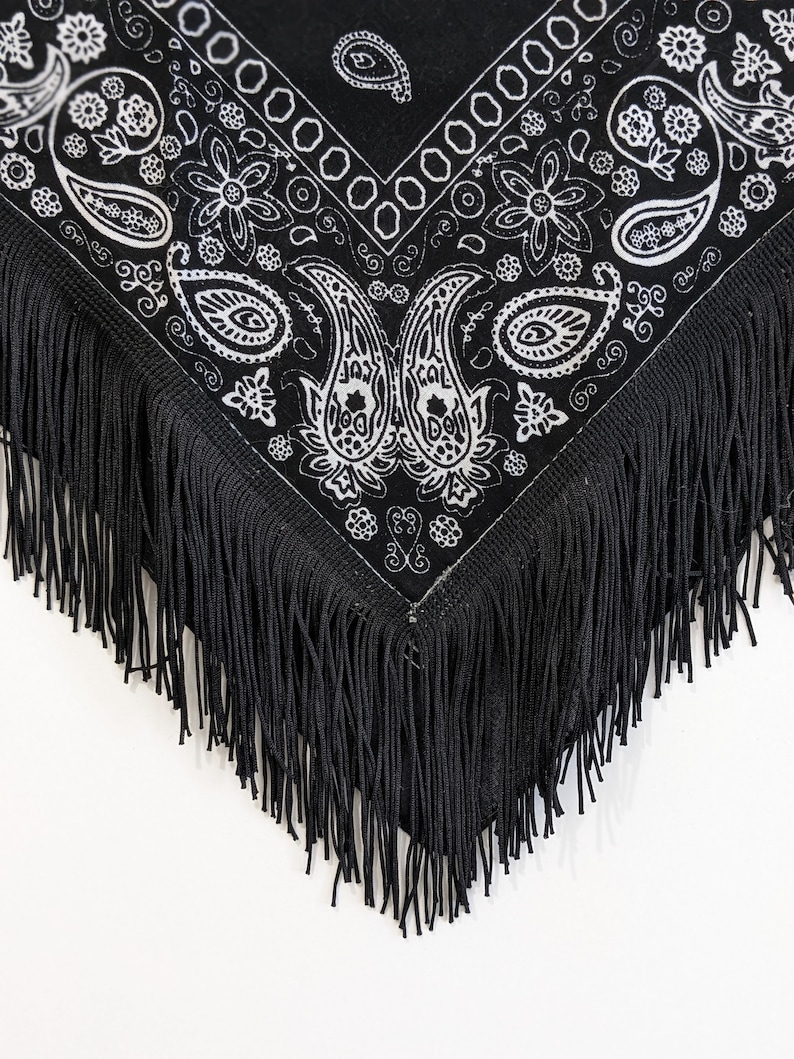 May include: A black triangular bandana featuring a white paisley and floral pattern printed across the fabric. The edges of the bandana are finished with long, thin black fringe that hangs down from the bottom point and sides of the garment.