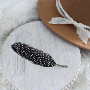 May include: A black and white speckled feather on a white wooden round tray with a brown felt hat with a white ribbon on top.