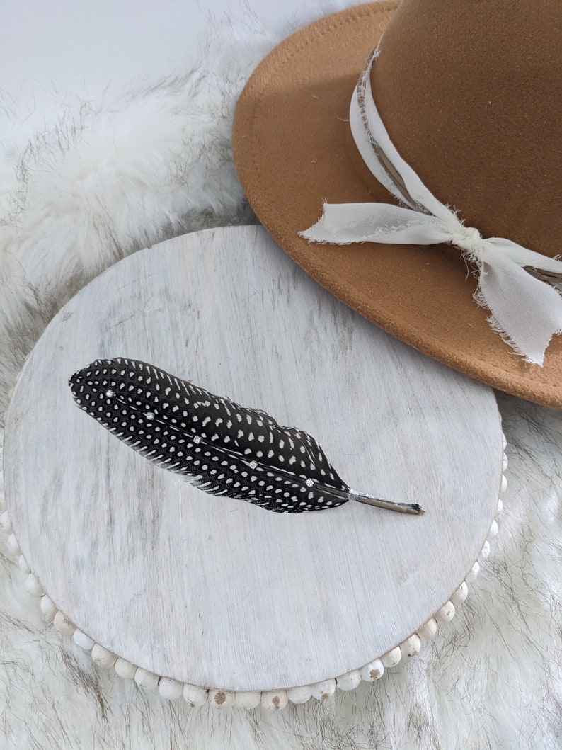 May include: A black and white speckled feather on a white wooden round tray with a brown felt hat with a white ribbon on top.