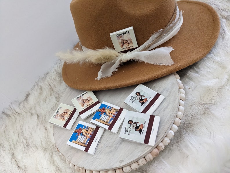 Pu&ograve; includere: Un cappello fedora marrone con un nastro bianco e una piccola scatola bianca con la scritta "Cowgirls" su di essa. Il cappello &egrave; appoggiato su una superficie bianca con cinque scatole di fiammiferi che mostrano immagini di donne in abiti da cowgirl. Le scatole di fiammiferi hanno la scritta "Cowgirls" e "Texas Sweethearts" su di esse.