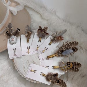 May include: A collection of seven lapel pins featuring assorted brown, grey, and tan feathers, dried floral accents, and metallic casings. One pin includes an oval silver-toned setting with a pink stone. Each pin is mounted on a white display card.