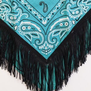 May include: A turquoise bandana featuring a white paisley pattern with intricate floral and teardrop motifs. The fabric is triangular in shape and is finished along the two bottom edges with a long, dense fringe made of thin black thread.