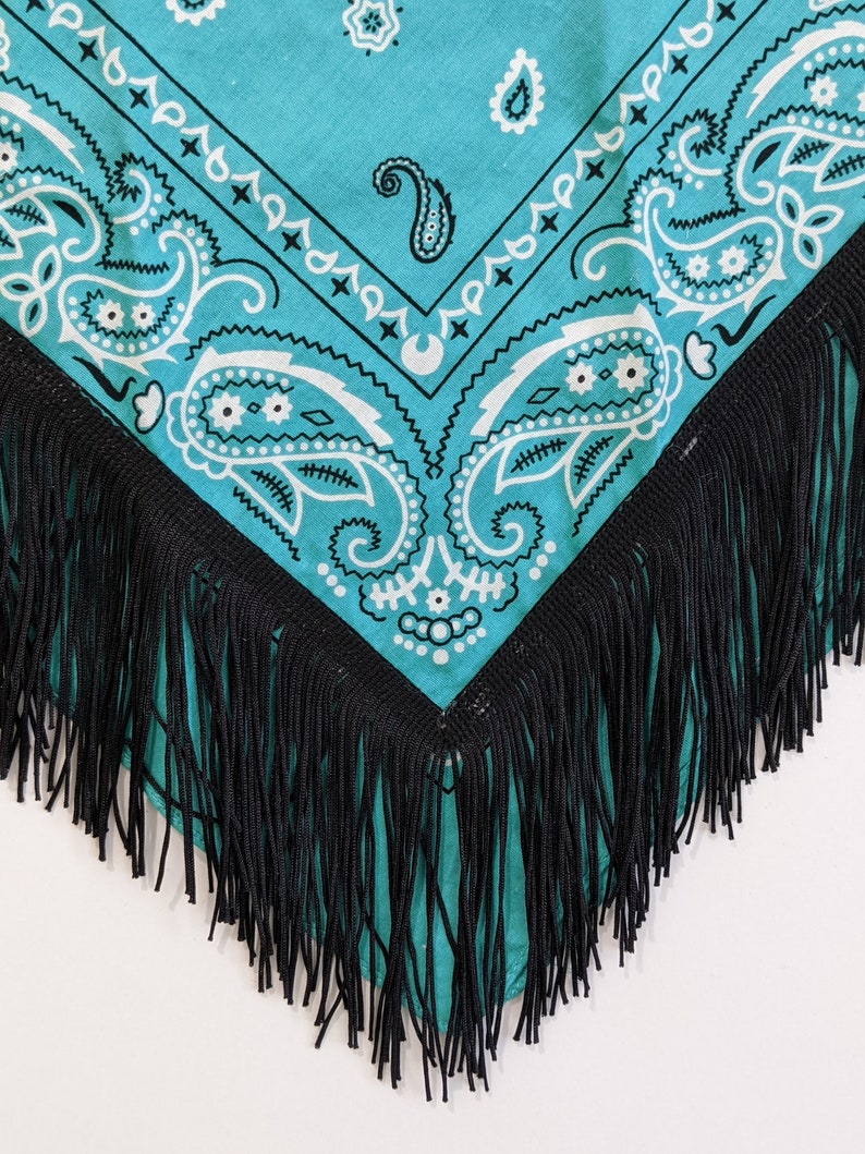 May include: A turquoise bandana featuring a white paisley pattern with intricate floral and teardrop motifs. The fabric is triangular in shape and is finished along the two bottom edges with a long, dense fringe made of thin black thread.