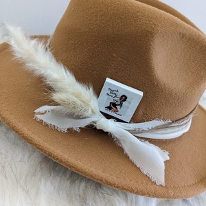 Pu&ograve; includere: Un cappello fedora in feltro marrone con una piuma bianca e un nastro bianco. Il cappello ha un piccolo adesivo quadrato bianco con il testo "Cowgirls Have More Fun" su di esso.