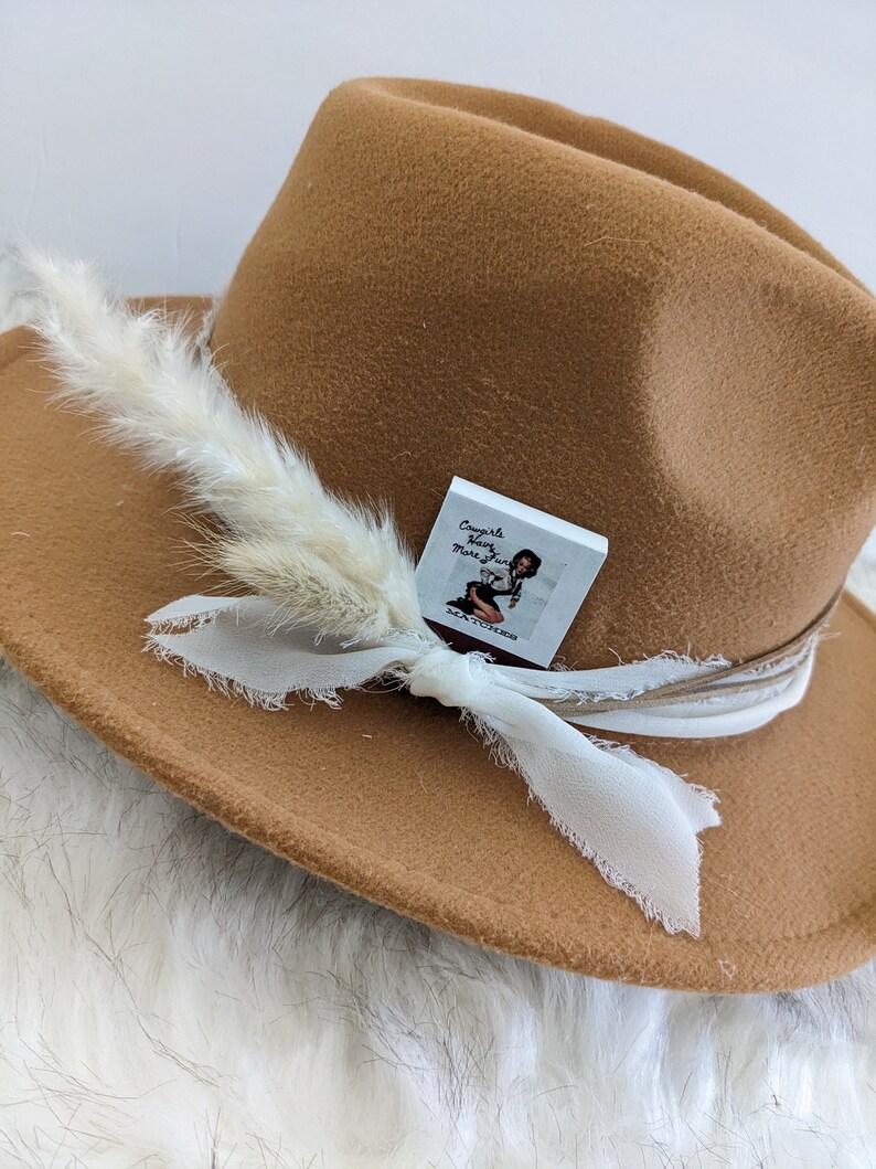 Pu&ograve; includere: Un cappello fedora in feltro marrone con una piuma bianca e un nastro bianco. Il cappello ha un piccolo adesivo quadrato bianco con il testo "Cowgirls Have More Fun" su di esso.