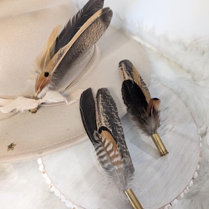 Może przedstawiać: Trzy brązowe i beżowe pióra z czarnymi końcówkami przymocowane do mosiężnych łusek pocisków. Pióra są ułożone na białym tle z brązowym kapeluszem w tle.