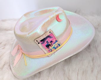 Könnte beinhalten: Schillernder Cowboyhut mit rosa Band und Spielkartenmuster. Der Hut hat einen rosa Mond und eine Spielkarte mit einem Cowboy auf einem Pferd. Der Hut hat ein gelbes Band und ein rosa Band.