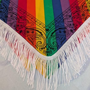 May include: A triangular bandana featuring vertical rainbow stripes in red, orange, yellow, green, blue, and purple. The fabric is decorated with a black paisley print along the edges and finished with long white fringe trim along the bottom border.