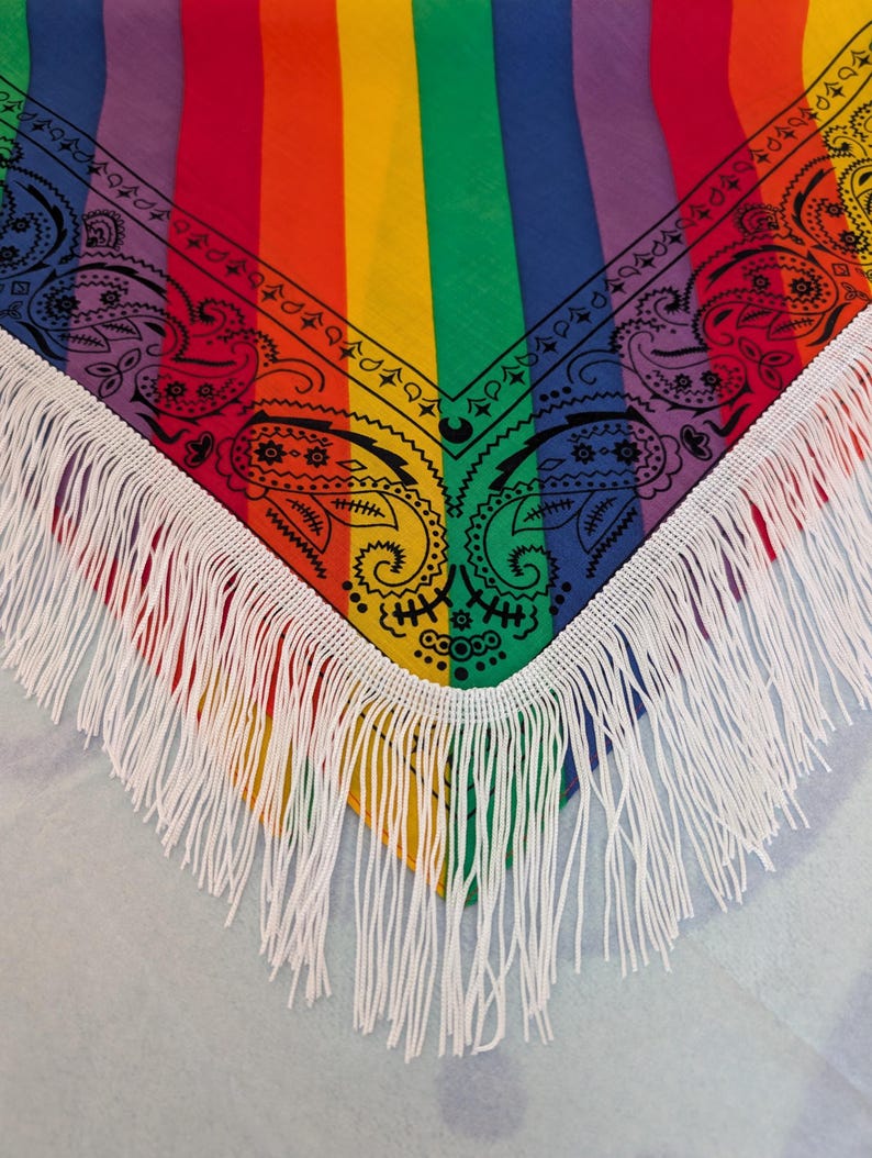 May include: A triangular bandana featuring vertical rainbow stripes in red, orange, yellow, green, blue, and purple. The fabric is decorated with a black paisley print along the edges and finished with long white fringe trim along the bottom border.