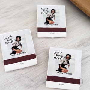 Pu&ograve; includere: Tre scatole di fiammiferi con sfondo bianco e una striscia marrone. Ogni scatola di fiammiferi presenta un'illustrazione in stile pin-up di una donna con un cappello da cowboy e un abito bianco e nero. Il testo su ogni scatola di fiammiferi dice "Cowgirls Have More Fun" e "MATCHES".