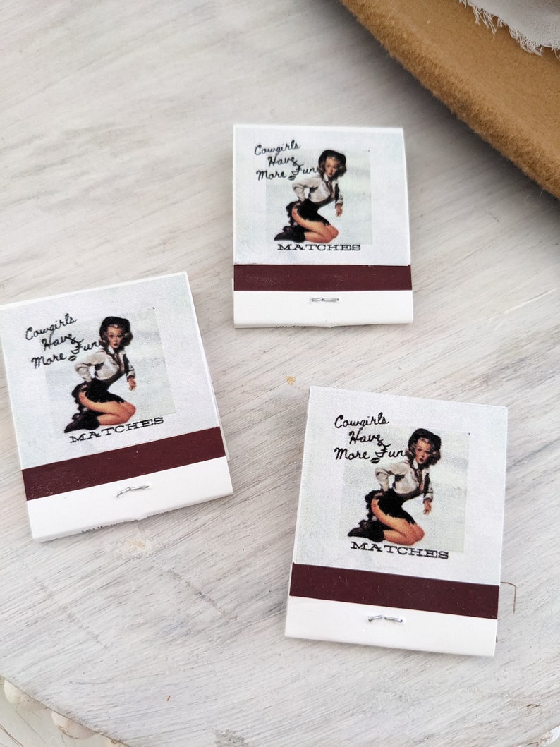 Pu&ograve; includere: Tre scatole di fiammiferi con sfondo bianco e una striscia marrone. Ogni scatola di fiammiferi presenta un'illustrazione in stile pin-up di una donna con un cappello da cowboy e un abito bianco e nero. Il testo su ogni scatola di fiammiferi dice "Cowgirls Have More Fun" e "MATCHES".