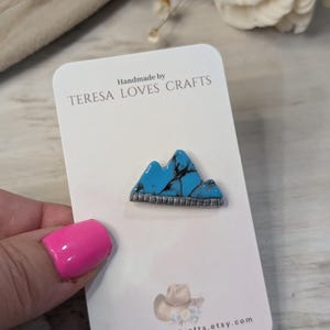 Może przedstawiać: Ręcznie robiony turkusowy wisiork w kształcie g&oacute;ry z srebrnymi akcentami na białej kartce z tekstem "Handmade by TERESA LOVES CRAFTS" i "TeresaLovesCrafts.etsy.com".