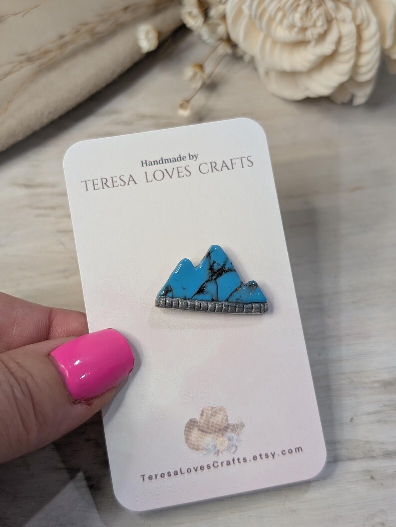Może przedstawiać: Ręcznie robiony turkusowy wisiork w kształcie g&oacute;ry z srebrnymi akcentami na białej kartce z tekstem "Handmade by TERESA LOVES CRAFTS" i "TeresaLovesCrafts.etsy.com".