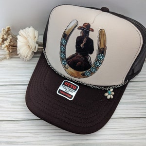 May include: A brown and beige trucker hat with a horseshoe and a cowgirl riding inside. The hat has a silver chain with a turquoise flower charm attached. The hat has a label that says "OTTO WORLD FAMOUS FOAM FRONT OS/FM TRUCKER".
