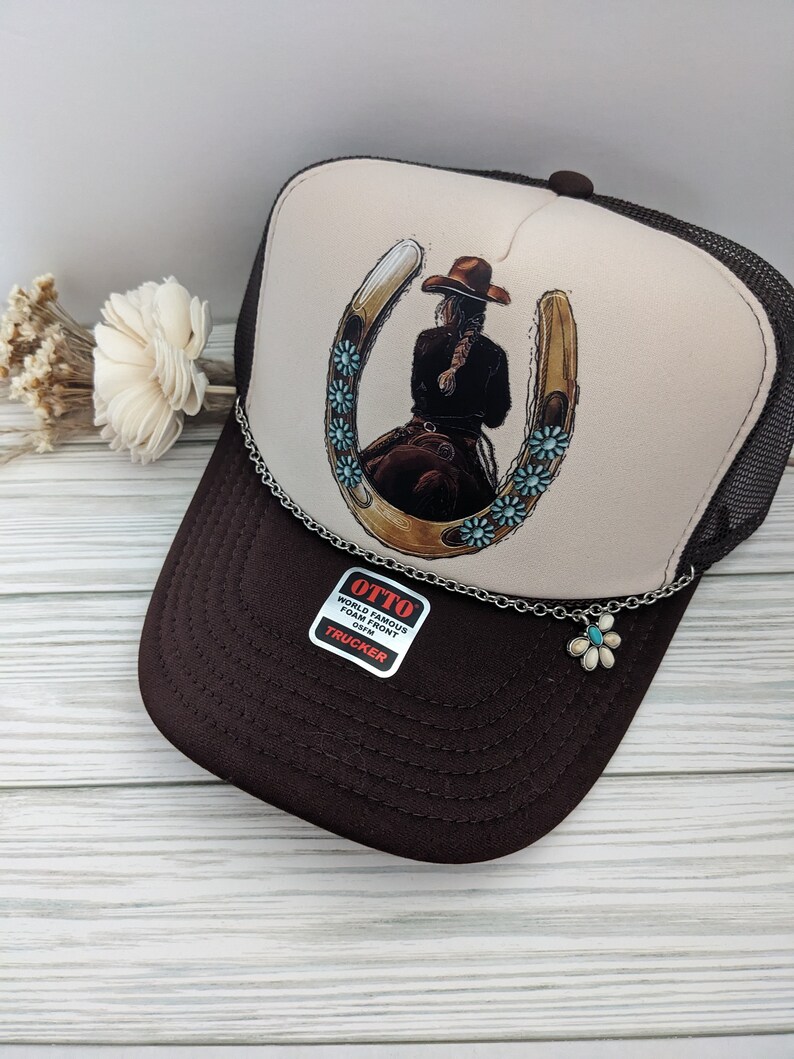 May include: A brown and beige trucker hat with a horseshoe and a cowgirl riding inside. The hat has a silver chain with a turquoise flower charm attached. The hat has a label that says "OTTO WORLD FAMOUS FOAM FRONT OS/FM TRUCKER".