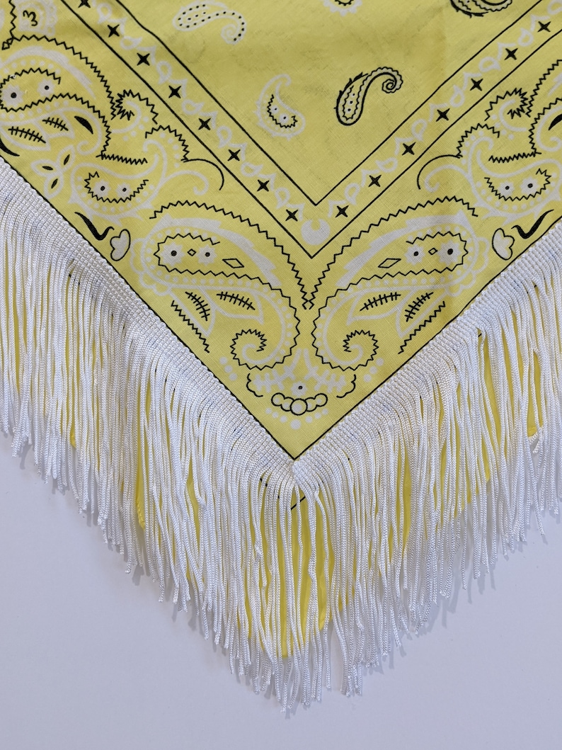 May include: A bright yellow bandana featuring a black paisley pattern and a white border. The fabric is finished with long, white fringe trim along the edges, creating a decorative accent on the triangular corner of the textile accessory.