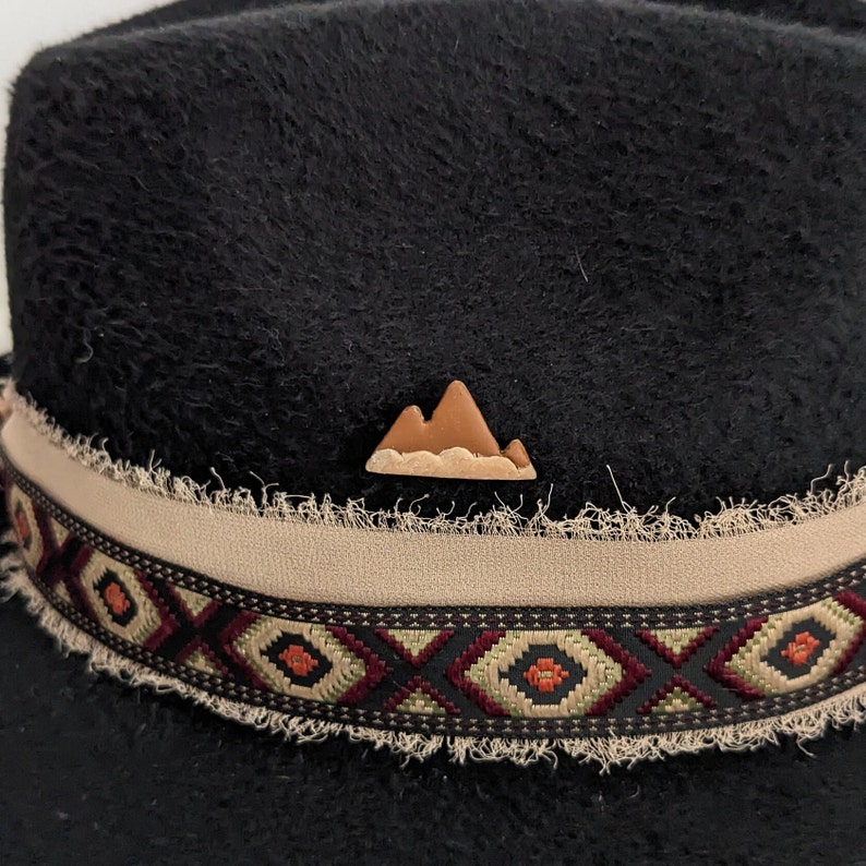 May include: Black felt fedora hat with a brown leather mountain pin and a beige and brown patterned band.