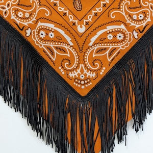 May include: A triangular bandana in a vibrant orange hue featuring a white paisley pattern. The edges of the fabric are adorned with long, thin black fringe trim that hangs down from the bottom point and sides of the garment accessory.