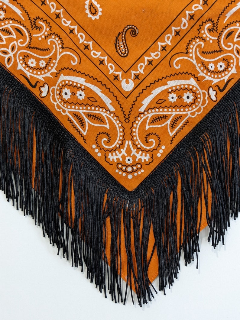 May include: A triangular bandana in a vibrant orange hue featuring a white paisley pattern. The edges of the fabric are adorned with long, thin black fringe trim that hangs down from the bottom point and sides of the garment accessory.