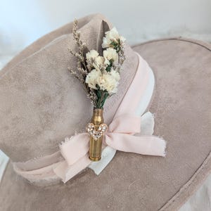 Può includere: Un cappello da cowboy beige con una composizione floreale decorativa. Il cappello presenta un nastro rosa chiaro e un piccolo bouquet di fiori bianchi in un bossolo di proiettile di ottone, adornato da un gioiello a forma di cuore.