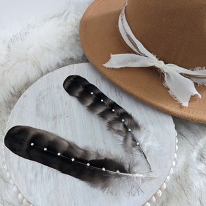 May include: Two black feathers with rhinestones on a white wooden round base with a brown fedora hat in the background.