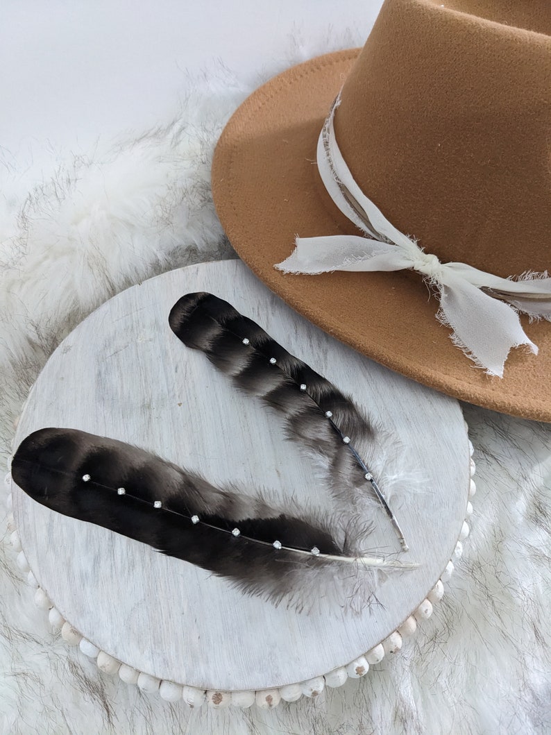 May include: Two black feathers with rhinestones on a white wooden round base with a brown fedora hat in the background.