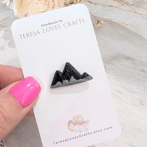 May include: Handmade by TERESA LOVES CRAFTS, a black and silver mountain range resin pin. The pin is on a white card with the shop's name and website address.