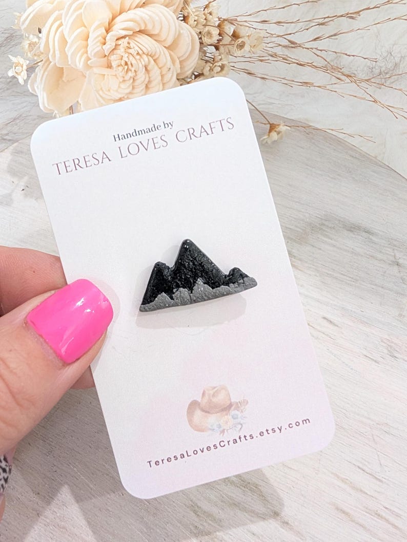 May include: Handmade by TERESA LOVES CRAFTS, a black and silver mountain range resin pin. The pin is on a white card with the shop's name and website address.
