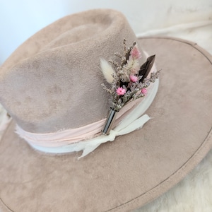 Può includere: Cappello fedora marrone chiaro con una composizione floreale decorativa. Il cappello presenta un nastro rosa e un nastro bianco, con un piccolo bouquet di fiori secchi e una piuma. Il cappello è realizzato in un materiale morbido, simile alla pelle scamosciata.