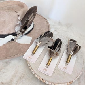 Può includere: Un cappello fedora color cammello con un dettaglio di piume e tre spille con piume. Le spille presentano varie piume attaccate a bossoli di proiettili in ottone, esposte su piccole carte bianche.
