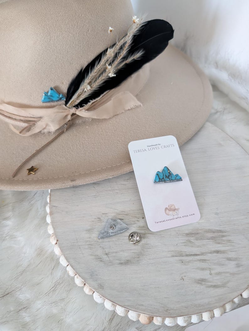 Może przedstawiać: Szpilka emaliowana przedstawiająca turkusowy pasmo g&oacute;rskie z srebrnym tyłem. Szpilka jest wyświetlana na białej kartce z tekstem "Handmade by TERESA LOVES CRAFTS" i adresem strony internetowej. Szpilka jest r&oacute;wnież pokazana na białym drewnianym podłożu z małym pi&oacute;rem i kapeluszem.
