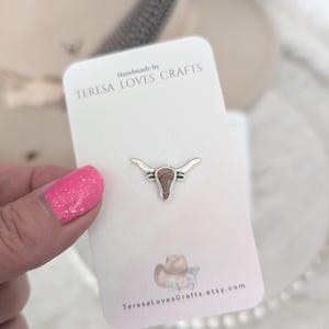 May include: A small silver-colored bull skull pin with a brown center is displayed on a white card. The card reads "Handmade by TERESA LOVES CRAFTS" and features a cowboy hat illustration. The background is blurred.