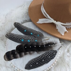 May include: Four black and white speckled feathers with turquoise accents arranged on a white beaded surface. A brown fedora hat with a white ribbon is in the background.