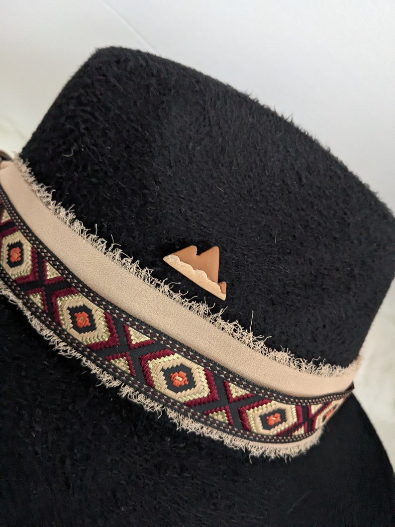 May include: Black felt fedora hat with a tan band featuring a geometric pattern. The band has a frayed edge and a small brown leather mountain pin.
