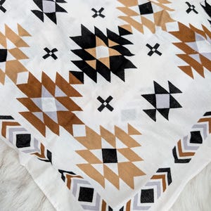 May include: A white fabric square featuring a repeating geometric pattern in shades of black, tan, and light gray. The design consists of interconnected diamonds, triangles, and small cross shapes arranged in a symmetrical, repeating layout.