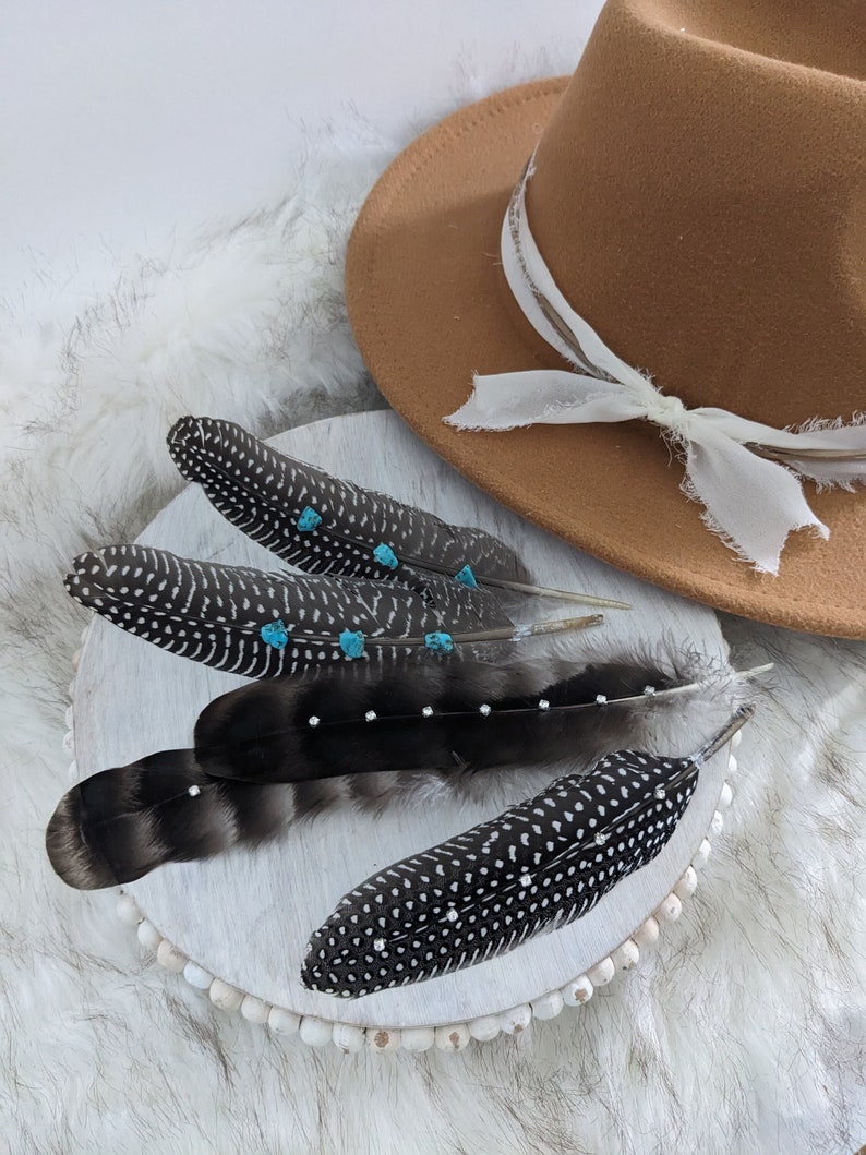 May include: Four black and white speckled feathers with turquoise accents on a white beaded surface. A brown fedora hat with a white ribbon is in the background.