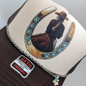 May include: A brown and white trucker hat with a horseshoe design featuring a woman riding a horse. The horseshoe is decorated with turquoise blue flowers. The hat has a chain with a turquoise blue flower charm attached.