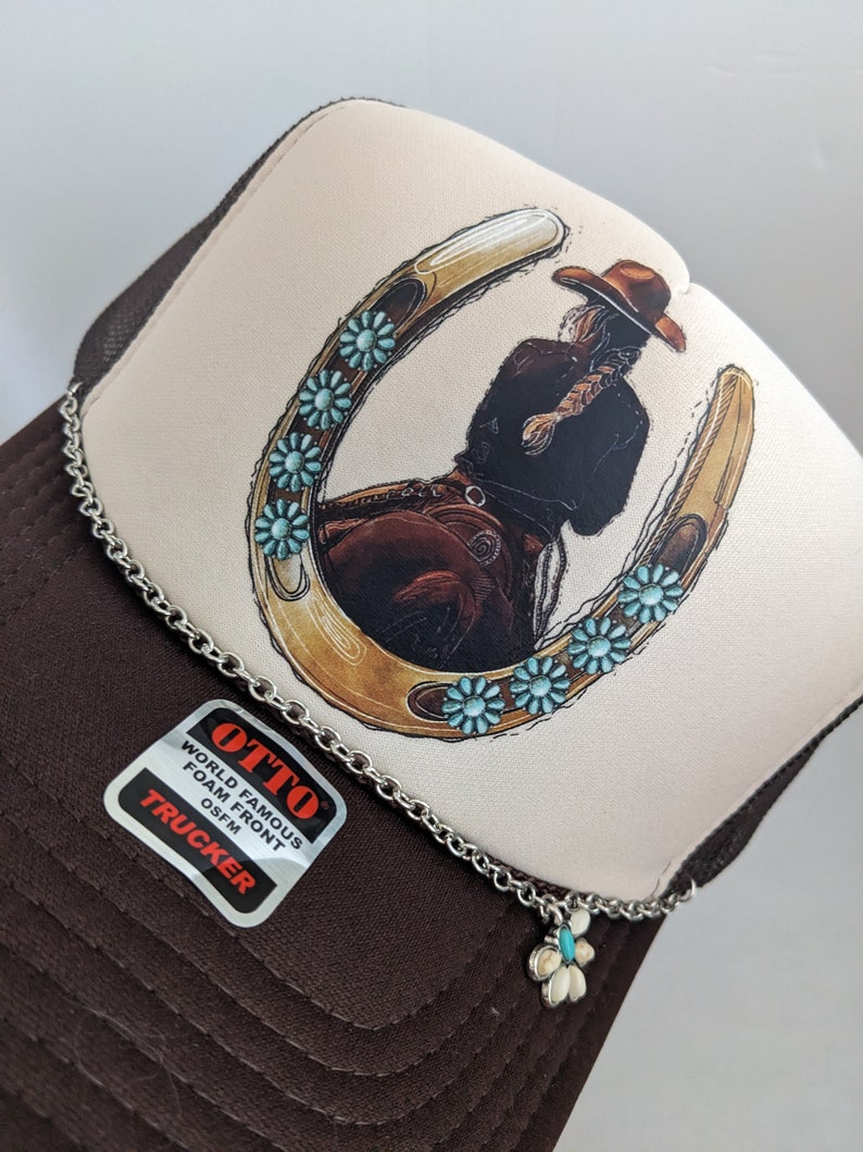 May include: A brown and white trucker hat with a horseshoe design featuring a woman riding a horse. The horseshoe is decorated with turquoise blue flowers. The hat has a chain with a turquoise blue flower charm attached.