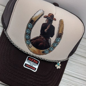 May include: A brown and white trucker hat with a brown mesh back. The front of the hat features a graphic of a woman riding a horse with a horseshoe around her. The horseshoe is decorated with turquoise blue flowers. The hat has a silver chain with a turquoise blue flower charm attached. The hat has a label that says "OTTO WORLD FAMOUS FOAM FRONT OSFM TRUCKER".