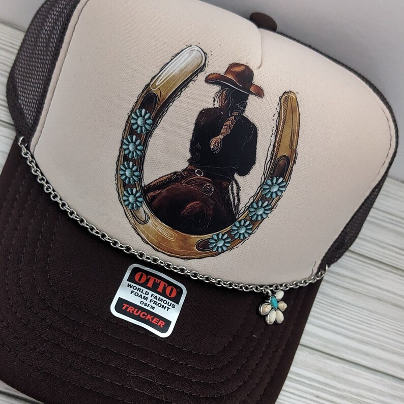 May include: A brown and white trucker hat with a brown mesh back. The front of the hat features a graphic of a woman riding a horse with a horseshoe around her. The horseshoe is decorated with turquoise blue flowers. The hat has a silver chain with a turquoise blue flower charm attached. The hat has a label that says "OTTO WORLD FAMOUS FOAM FRONT OSFM TRUCKER".