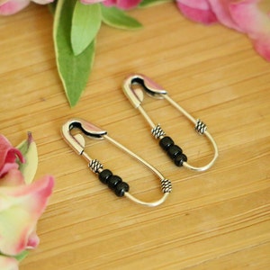 Peut inclure: Une paire de boucles d'oreilles en forme de punaise argentées avec des perles noires. Les boucles d'oreilles sont sur une surface en bois.