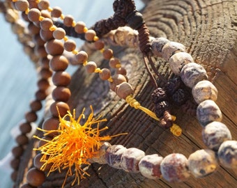 May include: Three strands of beaded necklaces with brown, gray, and black beads. The necklaces are tied together with a yellow cord and a small orange tassel. The necklaces are resting on a weathered wooden surface.