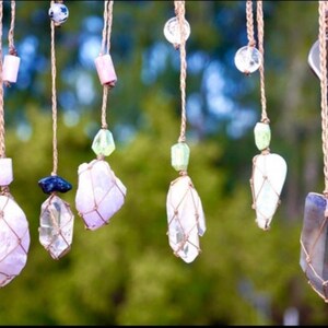May include: A collection of crystal pendants hanging from macrame string. The crystals are various shapes and sizes, including clear, pink, and blue. The macrame string is a natural brown color.