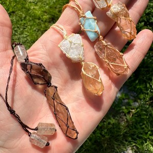 May include: Five crystal necklaces with different coloured stones wrapped in macrame. The stones are clear, brown, and light blue. The necklaces are made with brown and natural coloured cords.