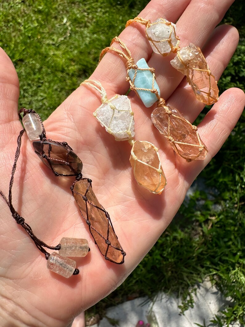 May include: Five crystal necklaces with different coloured stones wrapped in macrame. The stones are clear, brown, and light blue. The necklaces are made with brown and natural coloured cords.