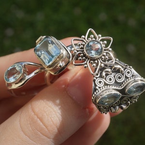 May include: Three silver rings with blue topaz gemstones. The rings feature intricate designs, including a flower, a swirl, and a rectangular setting.