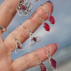 May include: A set of five pairs of silver earrings with ruby gemstones. The earrings feature different designs, including a circle with a ruby in the center, a teardrop shape with a ruby, and a Celtic knot with a ruby.